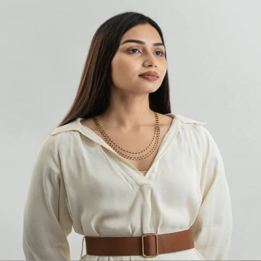 Woman wearing a white blouse with a brown belt and multiple necklaces on a plain background