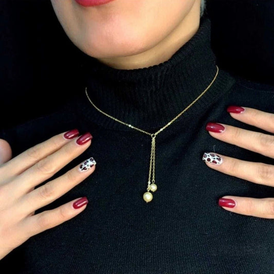 Person wearing a gold necklace with three pearl drops, hands with red nail polish and animal print design.