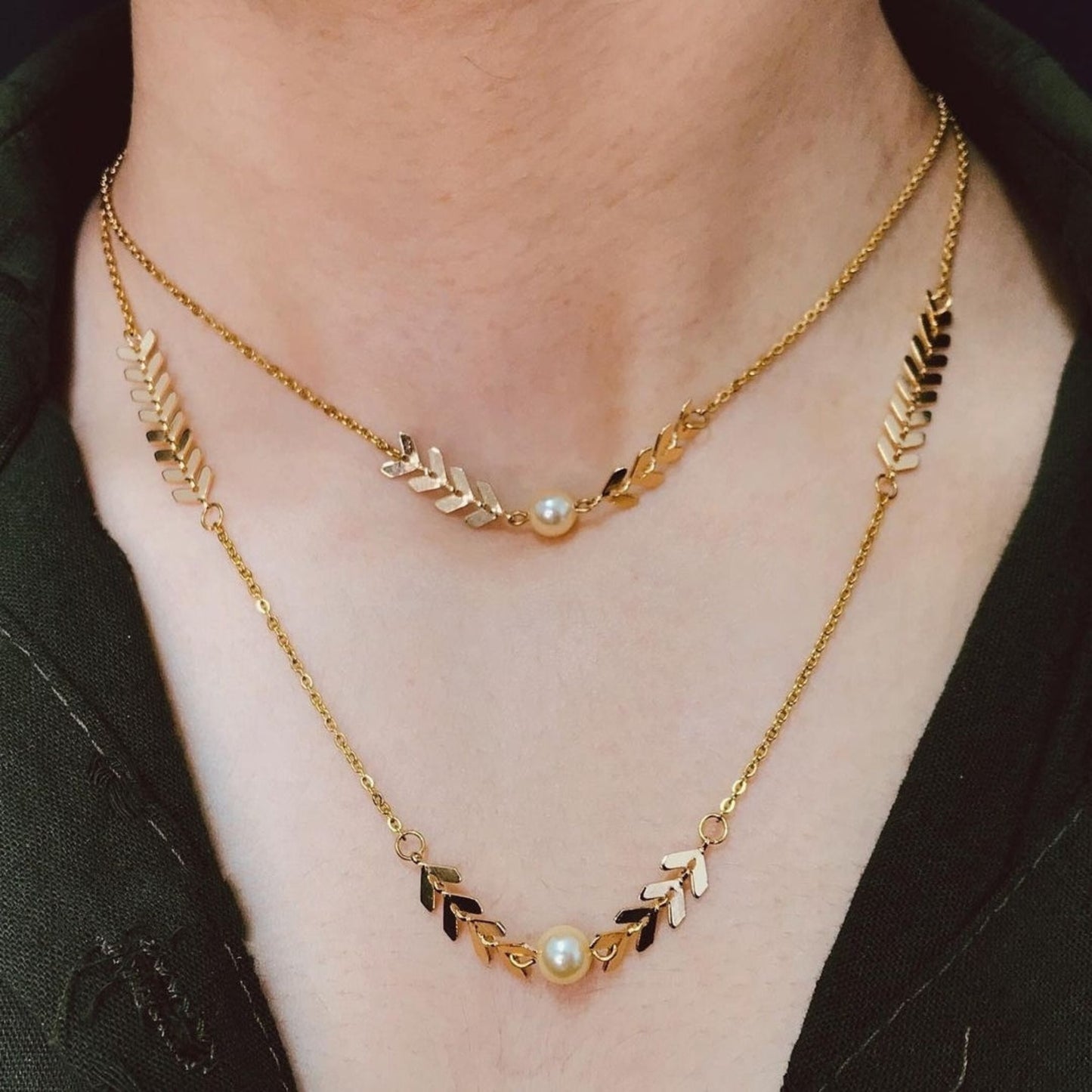 Two gold necklaces with leaf and pearl designs on a person wearing a black top.