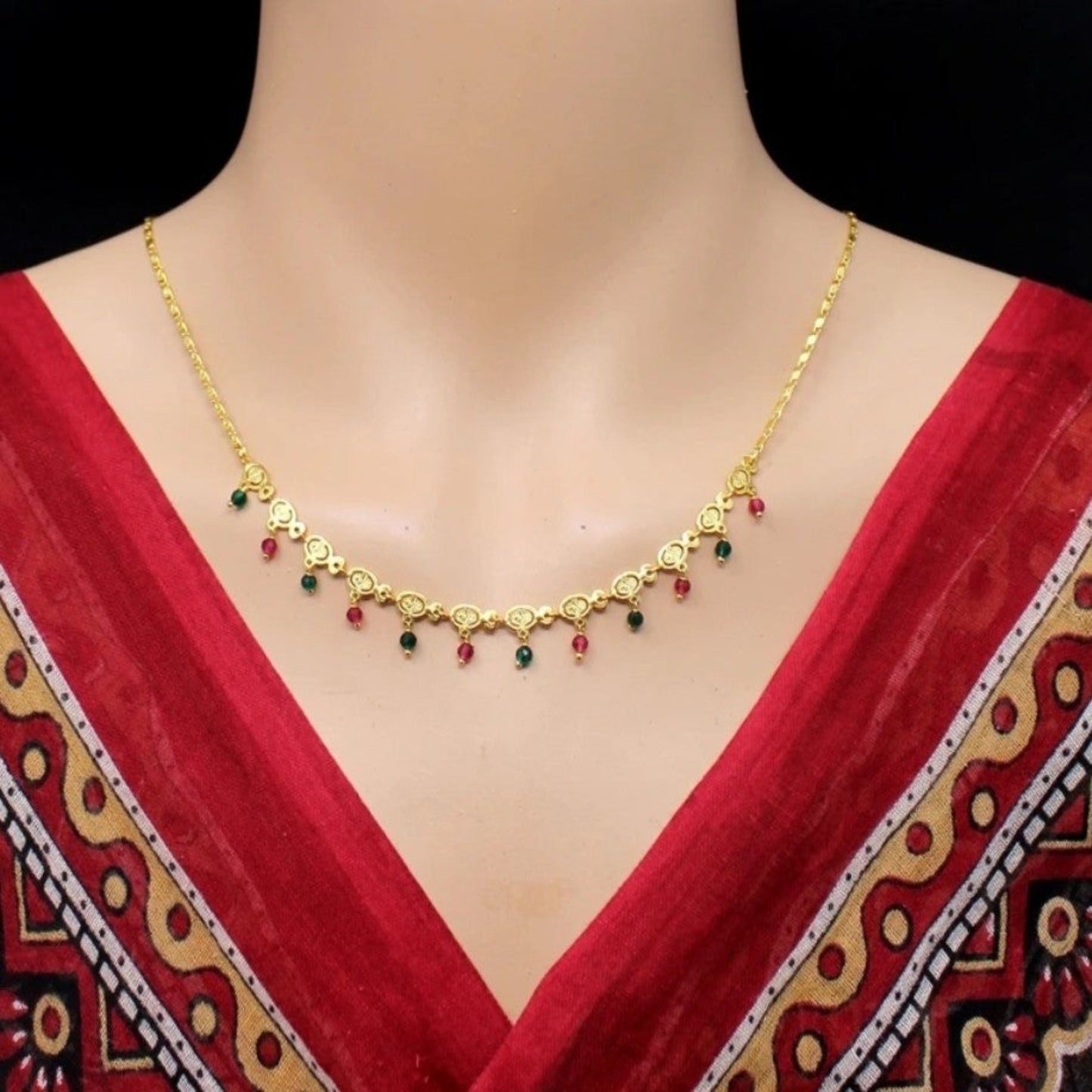 Gold necklace with red and green beads on a mannequin wearing a red saree.