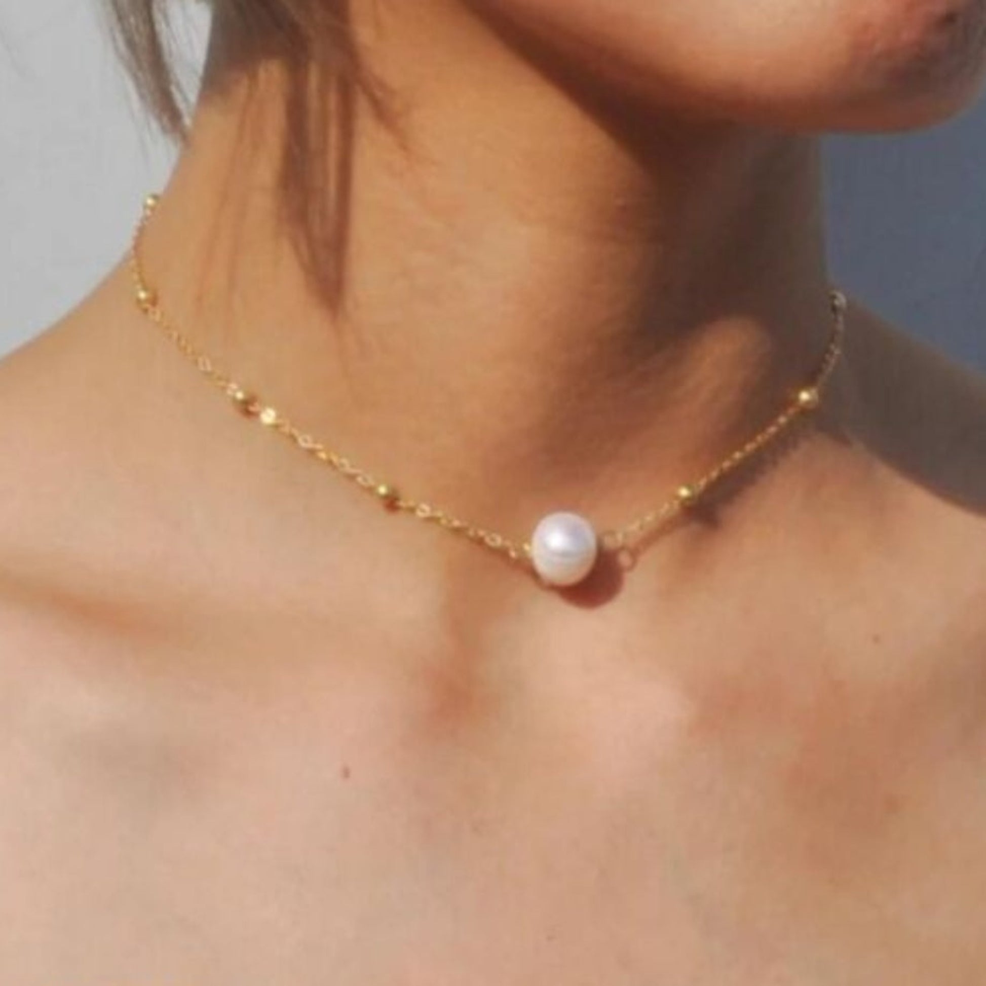 Gold necklace with a single pearl pendant on a person's neck against a neutral background