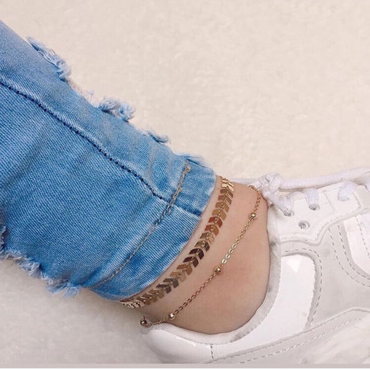Close-up of a person wearing gold anklets with white sneakers and blue jeans.