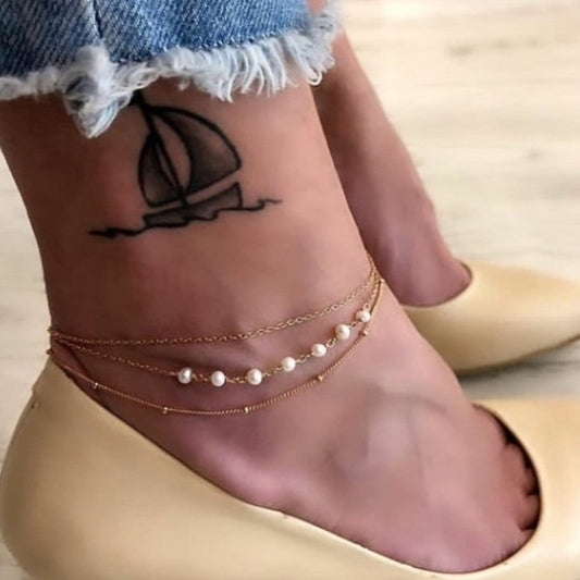 Close-up of a foot with multiple ankle bracelets and a sailboat tattoo on the ankle.