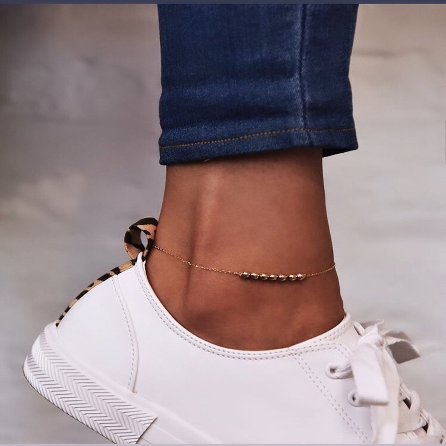 Close-up of a person's ankle with a gold anklet, wearing white sneakers and rolled-up jeans.