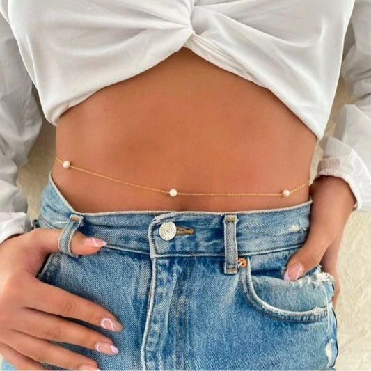 Person wearing a gold belly chain with small pearls, white top, and blue jeans.